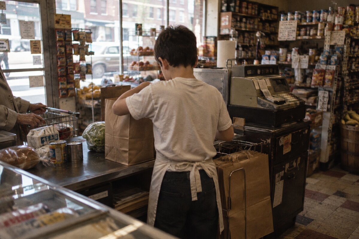 A boy in an apron bagging groceries at the register of a corner store